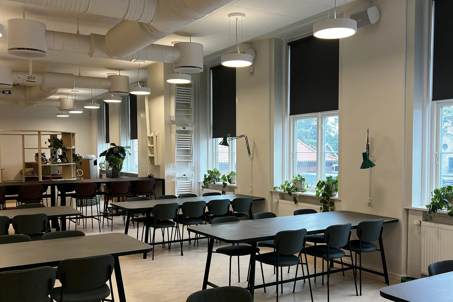 Fællesområde med mørke rullegardiner, grønne planter og arbejdsborde